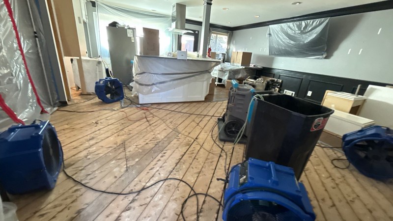 Blue air movers and an extraction unit running on a stripped plank subfloor behind plastic containment