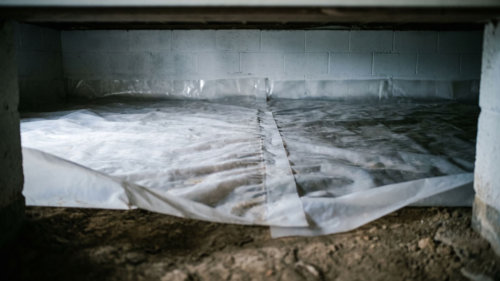 White polyethylene crawl space vapor barrier laid over bare soil with a lapped seam and the edge run up the block wall