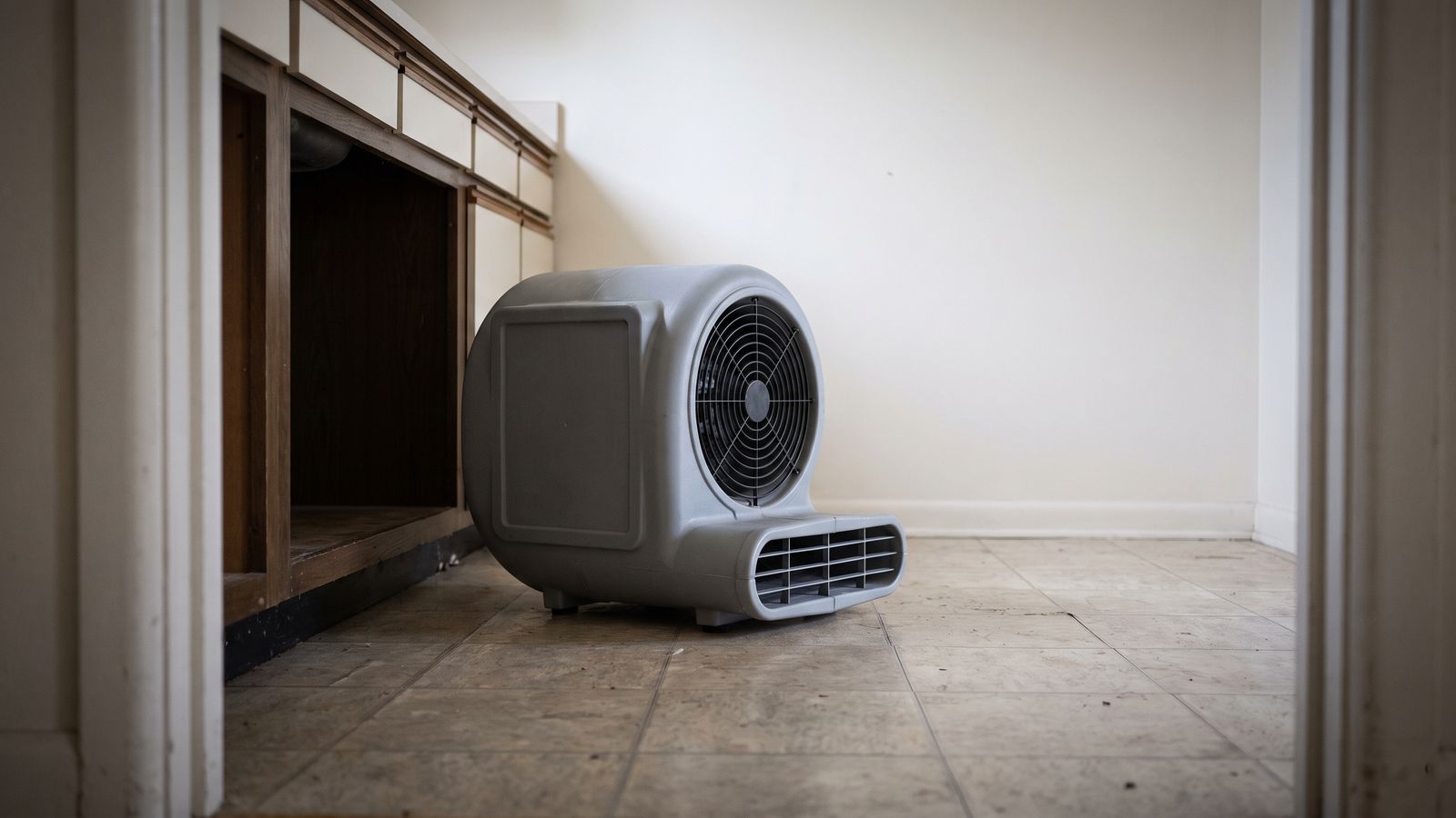 Compact centrifugal air mover on a tile floor beside the opened cabinet recess left by dishwasher leak water damage
