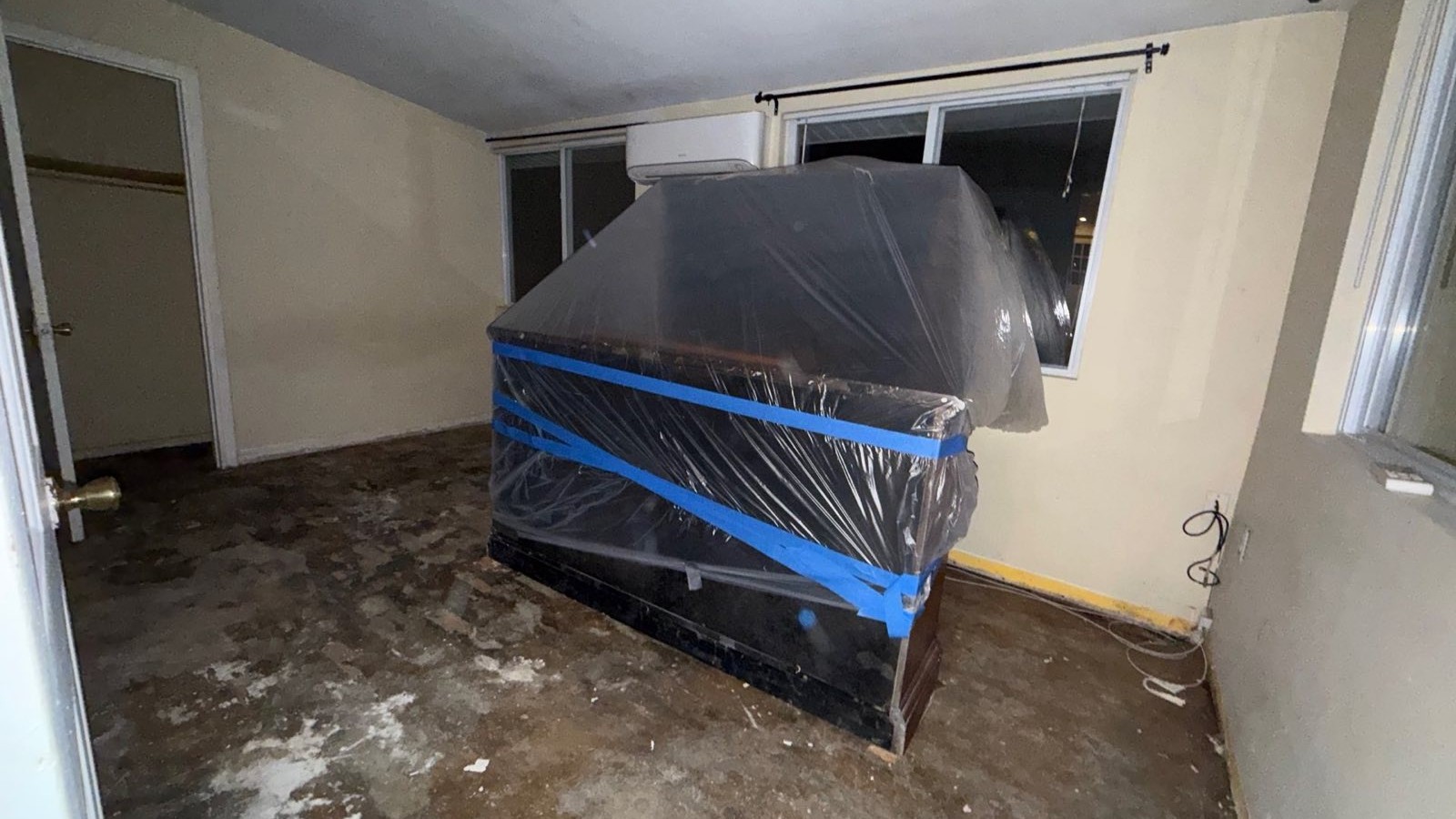 Furniture wrapped and taped in plastic in a room where the flooring has been torn down to the slab