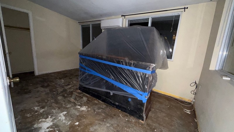 Furniture wrapped and taped in plastic in a room where the flooring has been torn down to the slab