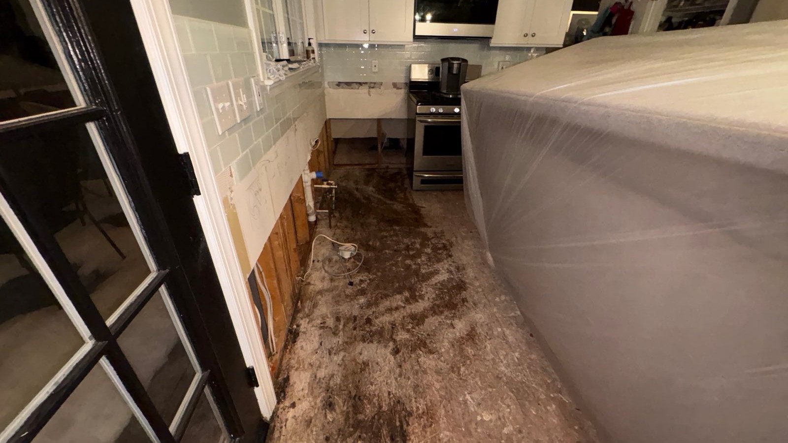 Kitchen flooring torn out to the subfloor with the lower wall opened and the island sealed in plastic