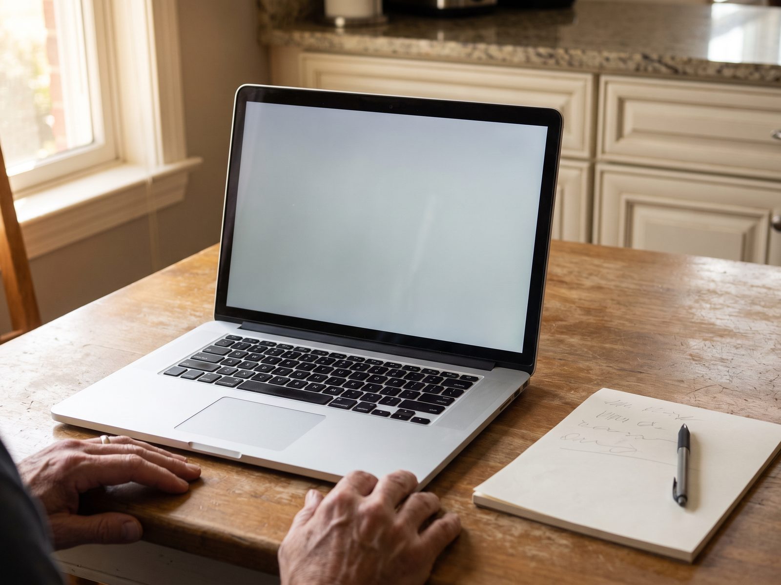Laptop with a blank screen open on a kitchen table beside a handwritten note and a pen.