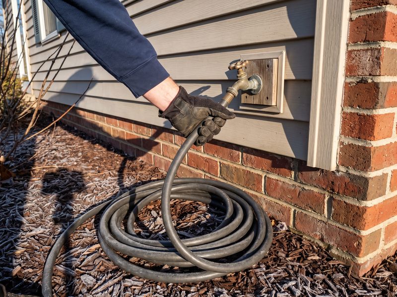Garden hose still attached to an outdoor spigot on a suburban house wall on a frosty morning as a gloved hand reaches to uncouple it.