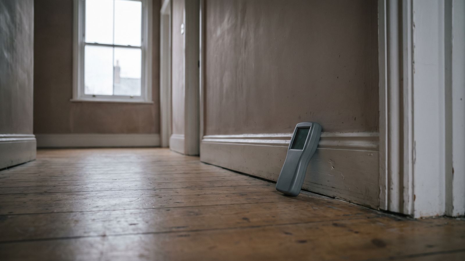 Moisture meter at a hallway baseboard Gray pinless moisture meter leaning against the baseboard of an empty hallway with worn oak floorboards and a sash window.
