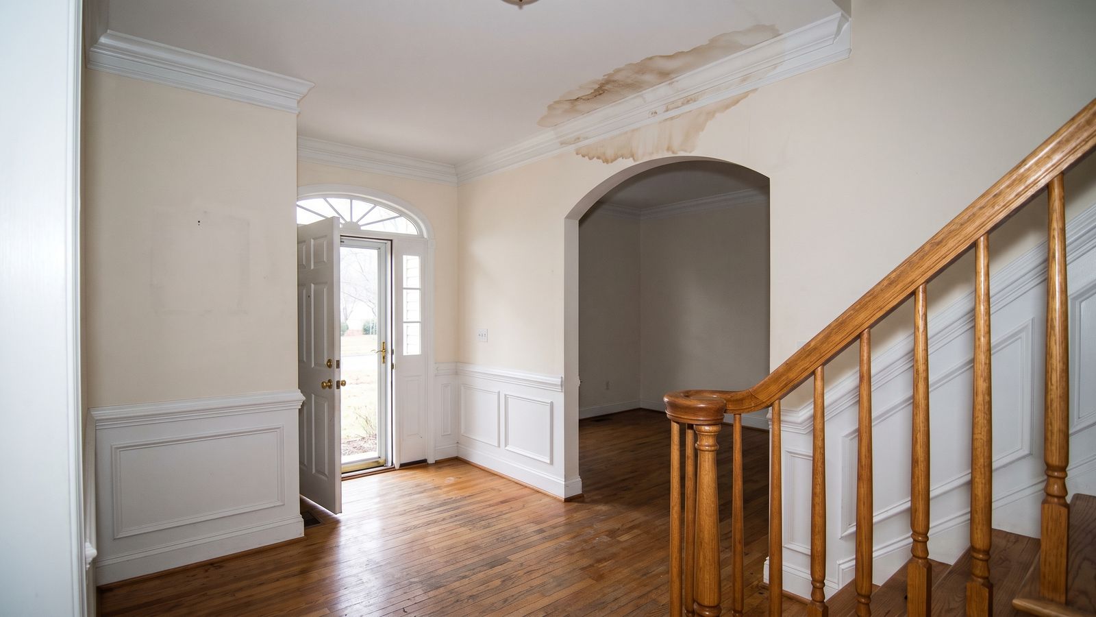Brown water stain spreading across the ceiling of an empty colonial entry hall, front door open beside the oak stair rail.