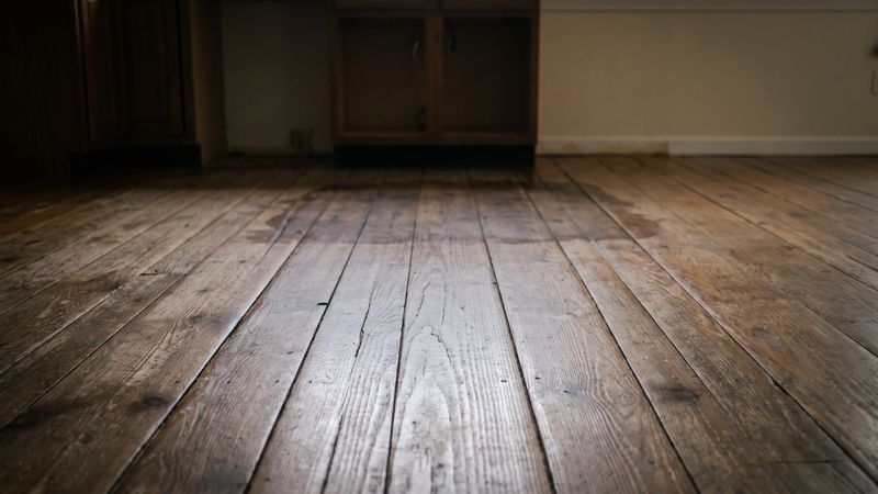 Dark damp stain across plank flooring at a kitchen cabinet base, early refrigerator water line leak floor damage