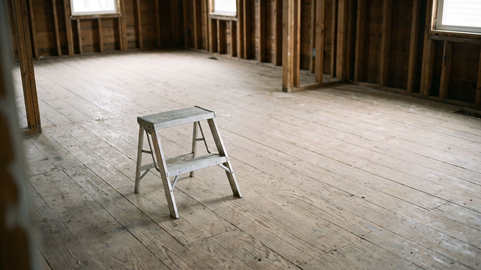 An emptied room between trades A folding aluminum step stool standing alone on a bare plank subfloor in an emptied wood-framed room.