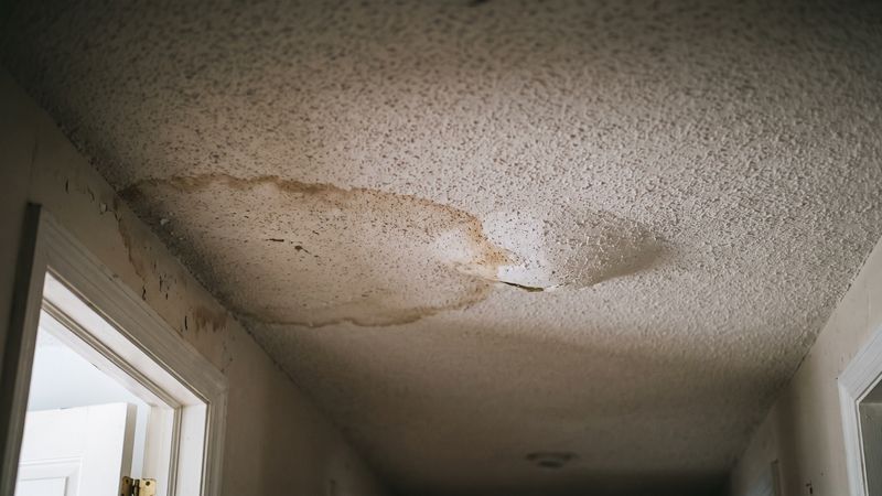 Brown tide stain and a torn patch overhead in an upstairs hallway, a water damage popcorn ceiling asbestos warning sign