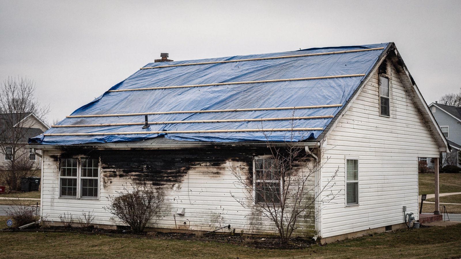 Blue poly tarp battened over a fire-damaged roof slope above soot-blackened siding after a house fire