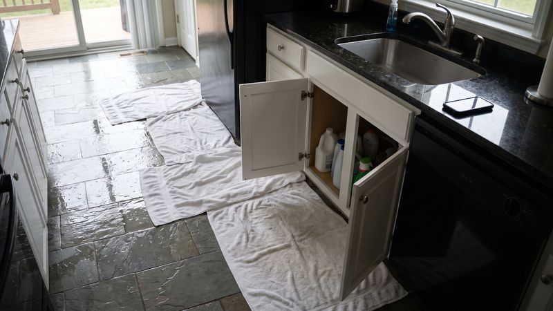 Kitchen floor toweled after a water leak, the sink base cabinet open and a phone resting screen-down on the counter.
