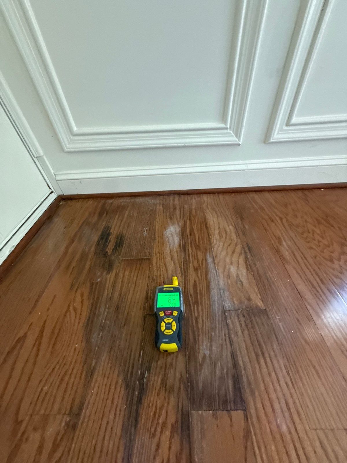 Handheld moisture and humidity meter lying on oak floor boards beside white wainscot, Manassas, VA, April 2026