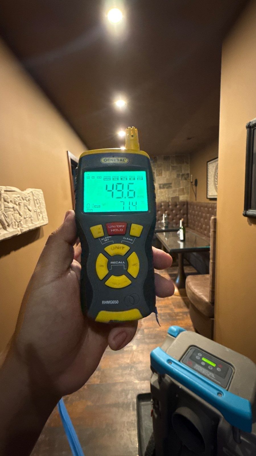 Handheld meter reading 49.6 percent relative humidity in a basement hallway with a stone accent wall and a dehumidifier, McLean, VA, October 25, 2025