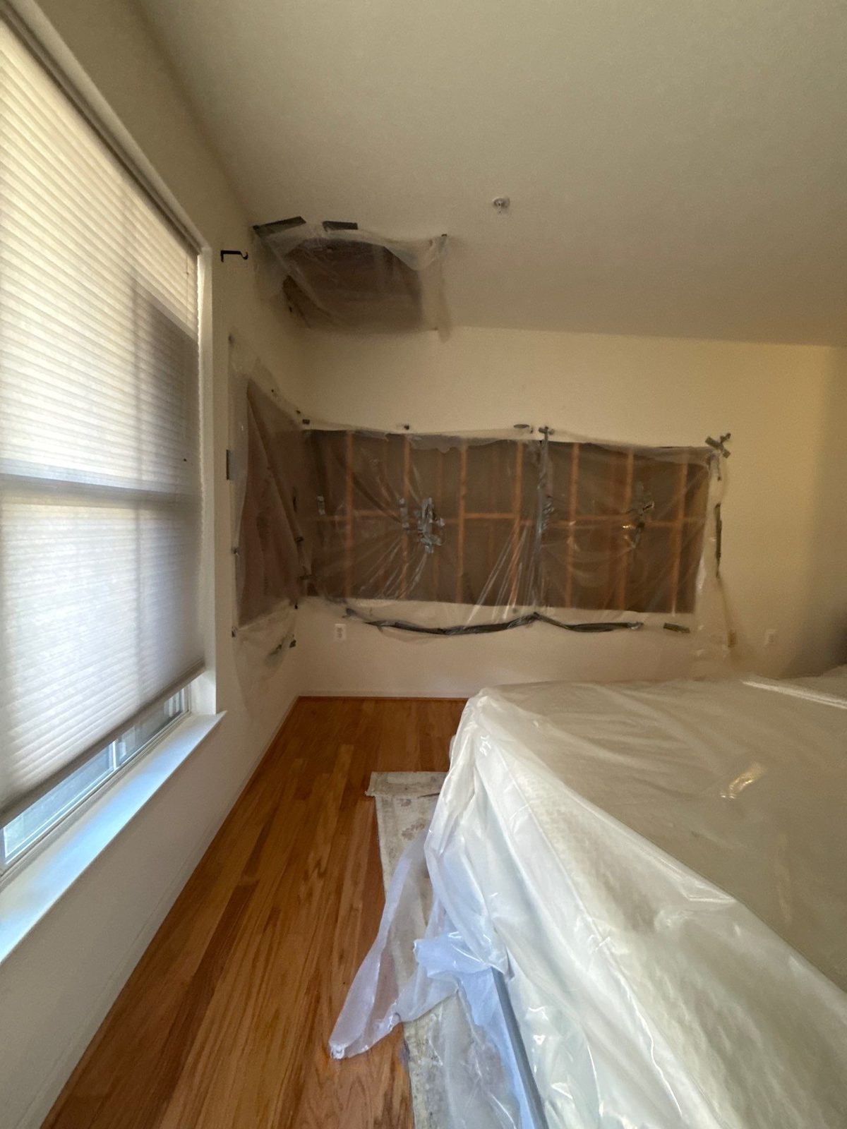 Opened bedroom wall under taped plastic beside a window, with a bed wrapped in plastic, Alexandria, VA, November 2025