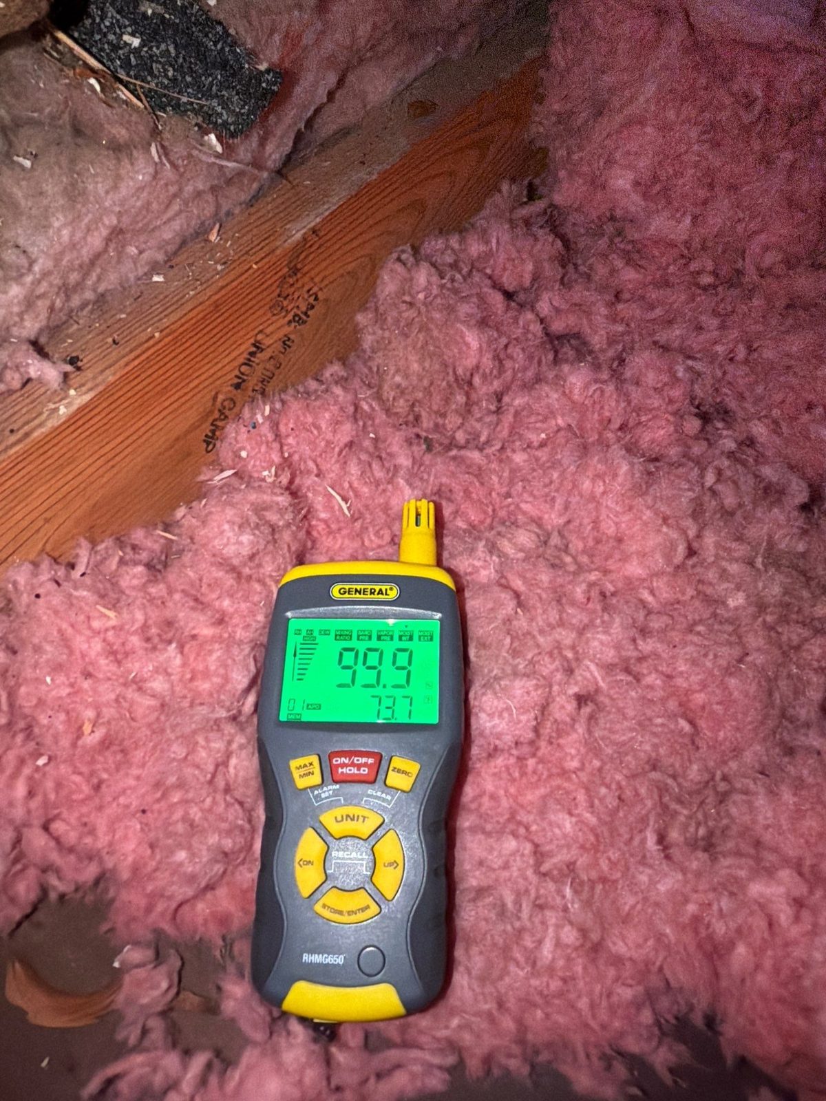 Handheld meter lying on pink fiberglass insulation next to a joist in an attic, Vienna, VA, March 2026