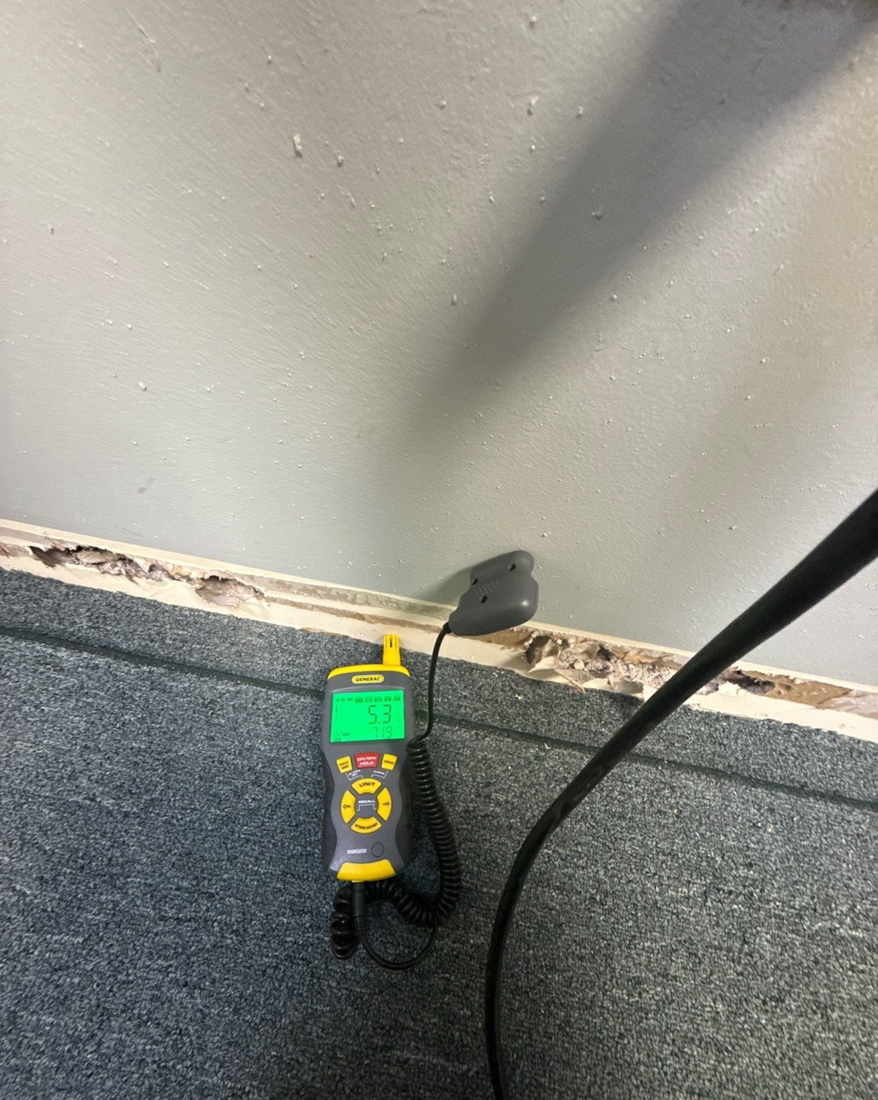 Moisture meter on carpet with its probe lead running to a wall base where the baseboard is chipped, reading 5.3, Sterling, VA, September 22, 2025
